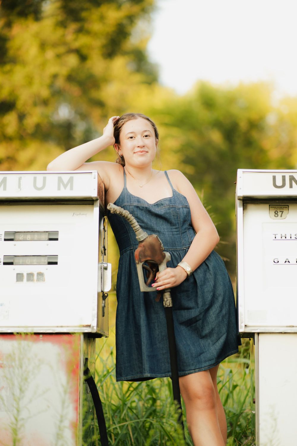 Young woman in denim dress posing between vintage gas pumps holding a fuel nozzle outdoors.