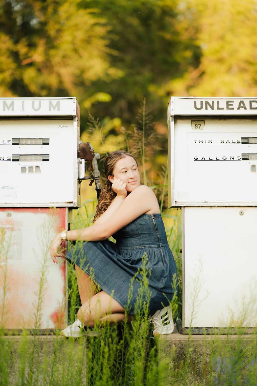 Young woman in denim overalls posing between vintage gas pumps surrounded by tall green grass and golden foliage.