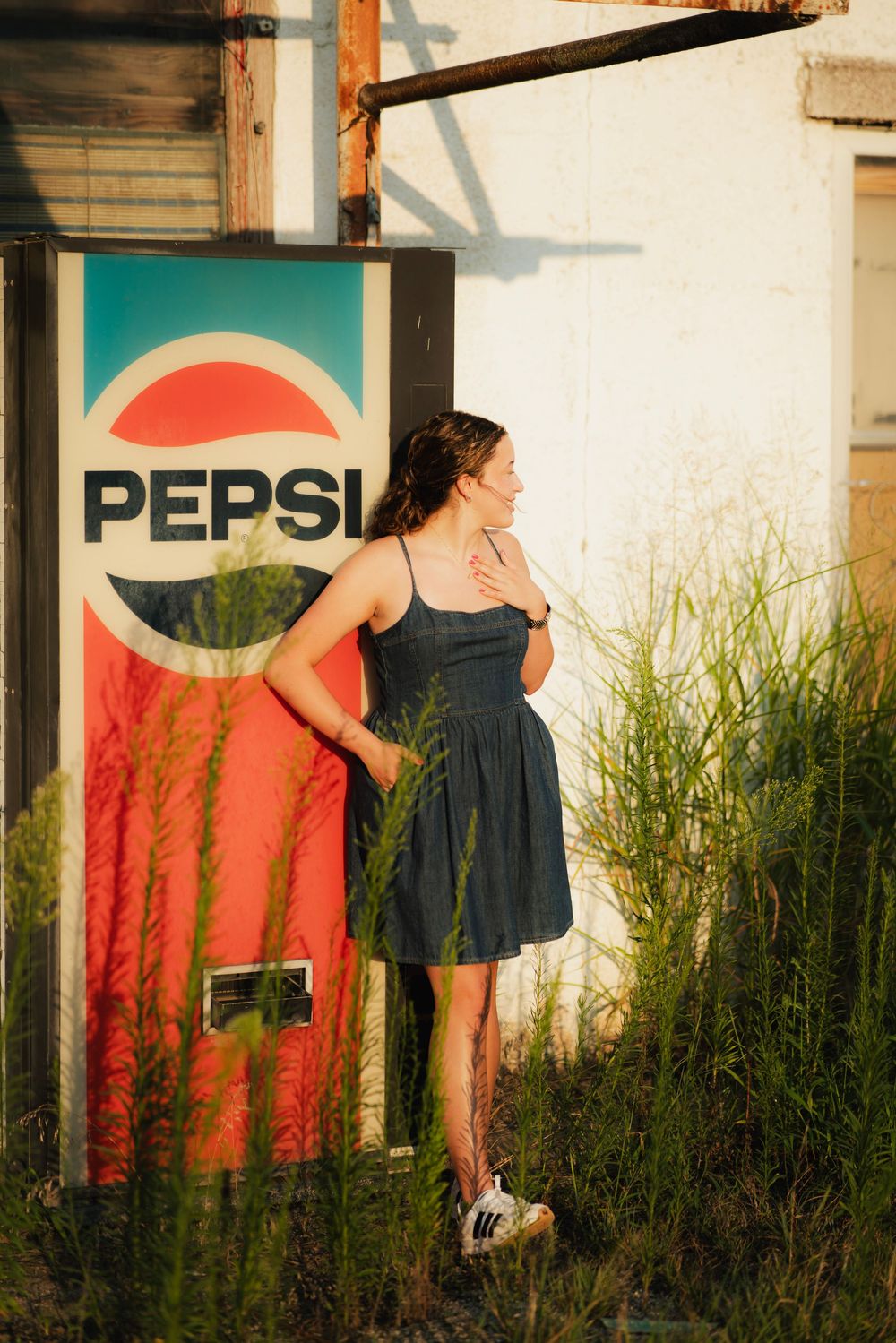 Young woman in blue dress standing by vintage Pepsi vending machine surrounded by tall green grass in golden hour light.