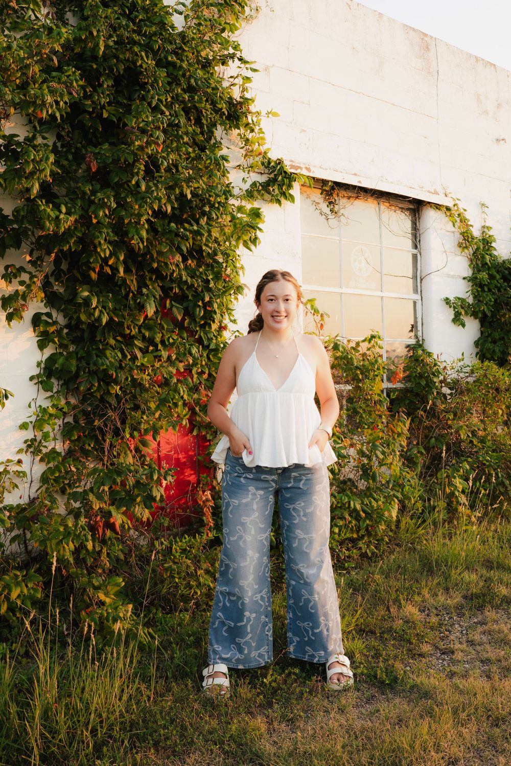 Young woman smiling in white halter top and floral jeans outdoors near ivy-covered white building in golden hour light.