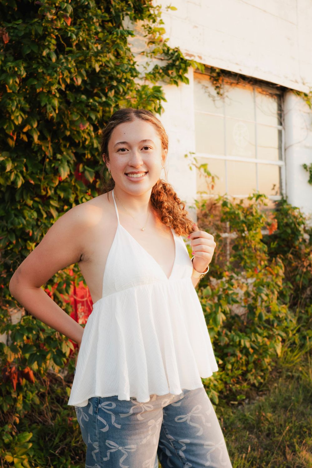 Smiling young woman in white flowy halter top and patterned jeans posing outdoors near ivy-covered wall in warm sunlight.