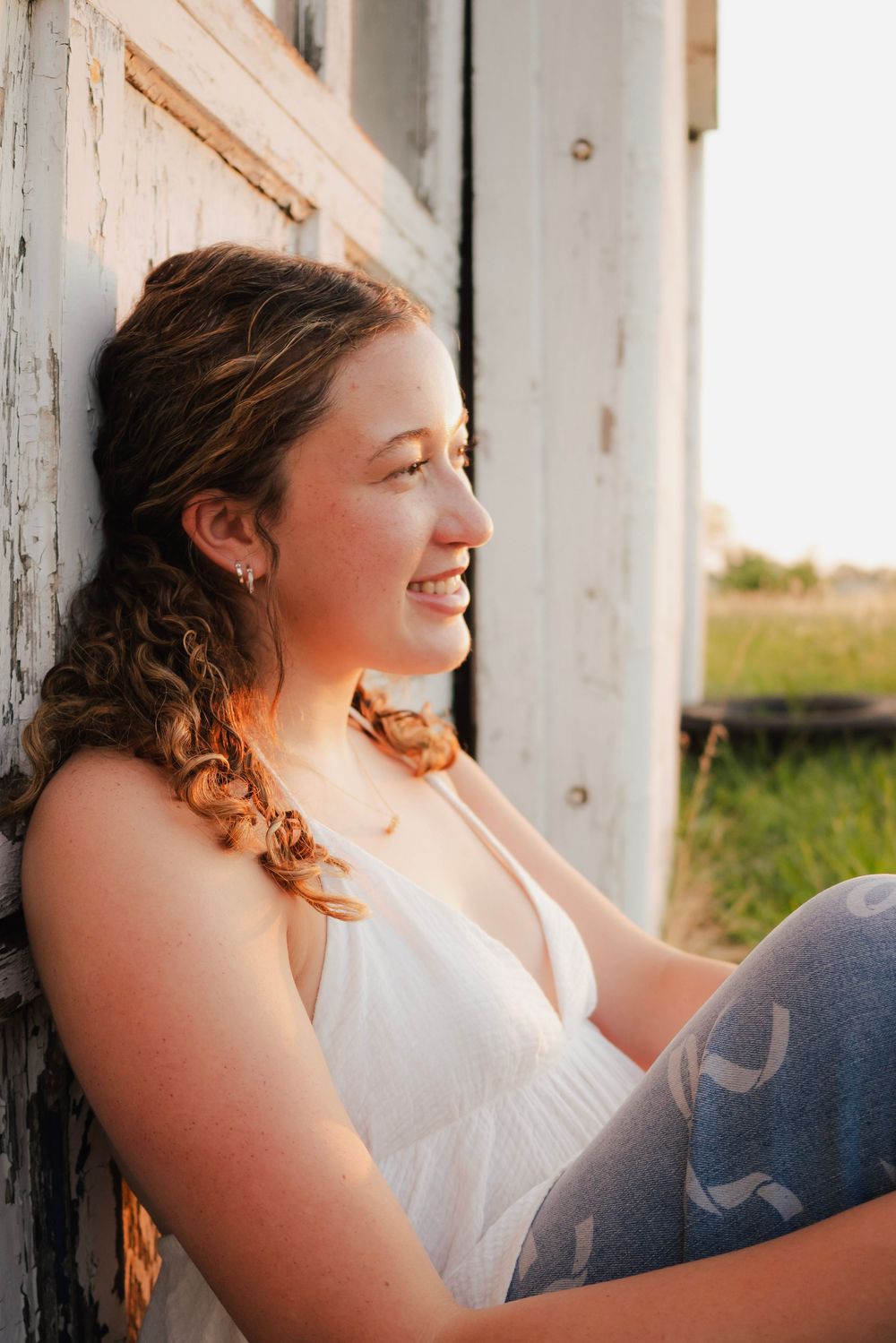 Young woman with curly hair smiles while leaning against a rustic white barn wall in golden hour sunlight.