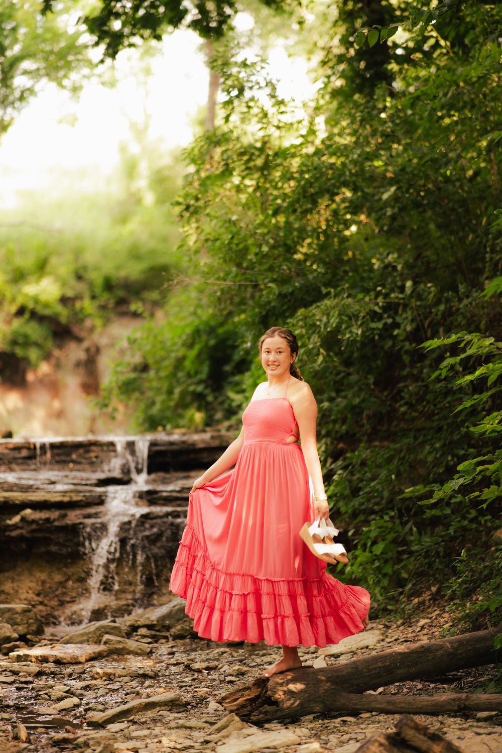 Young woman in coral ruffled midi dress holding shoes near a waterfall surrounded by lush greenery.
