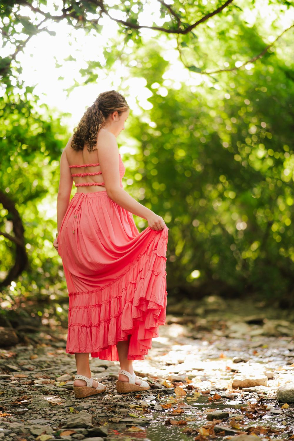 Young woman in pink ruffled dress lifting skirt while standing on rocky creek bed surrounded by lush green trees.