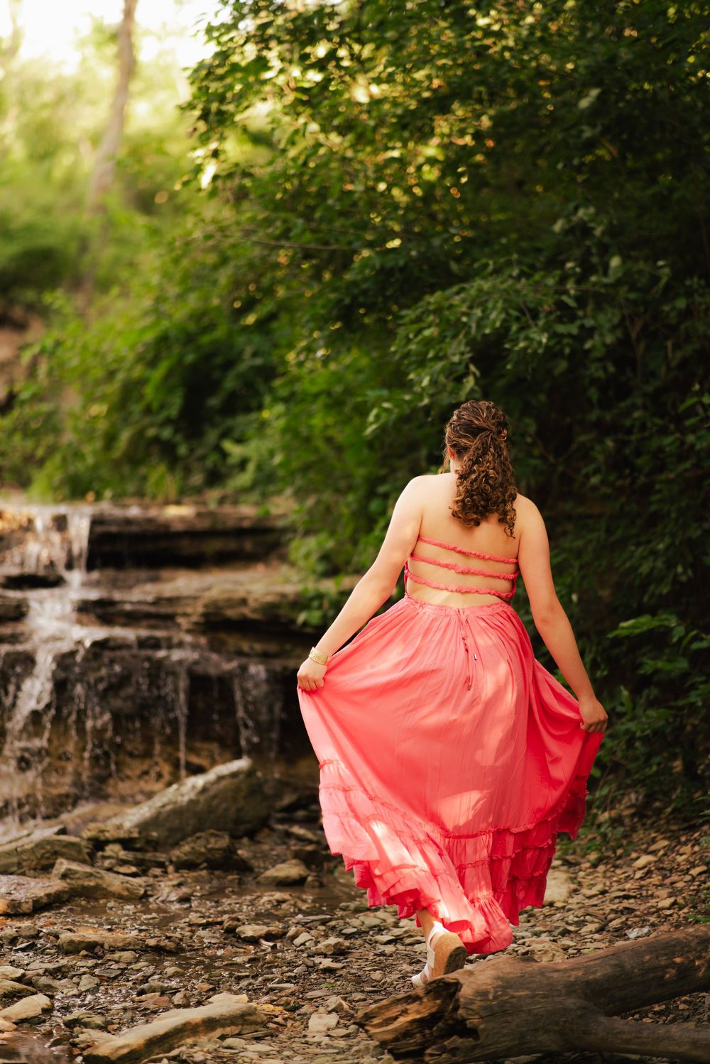 Woman in coral pink two-piece dress walks toward a woodland waterfall, holding her flowing skirt on rocky terrain.