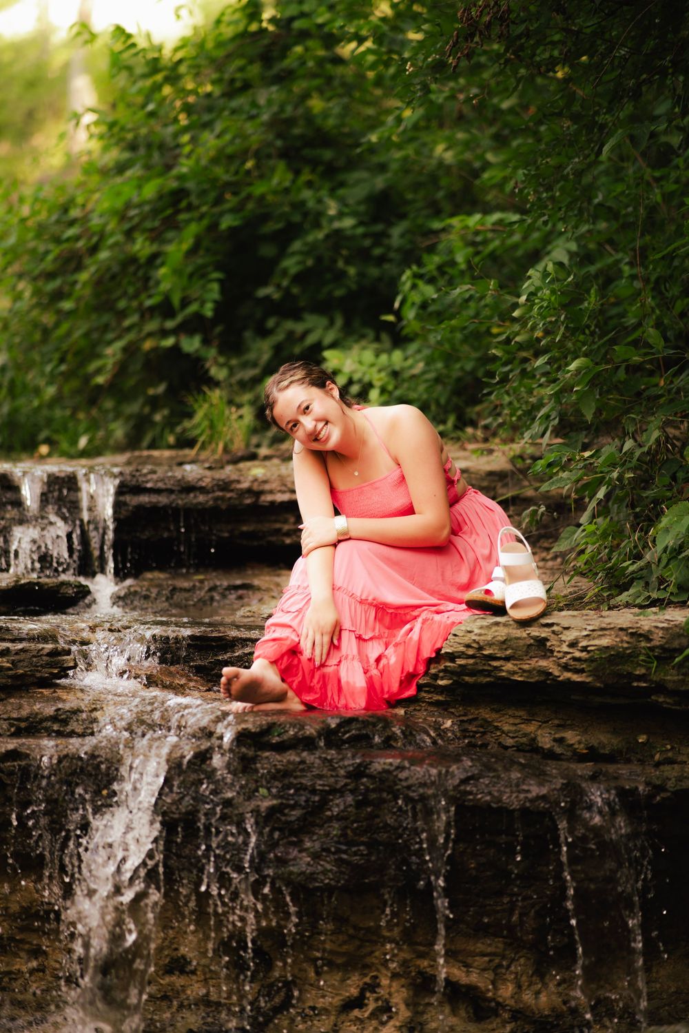 Smiling young woman in coral pink dress sitting barefoot on rocks beside a small waterfall in nature.