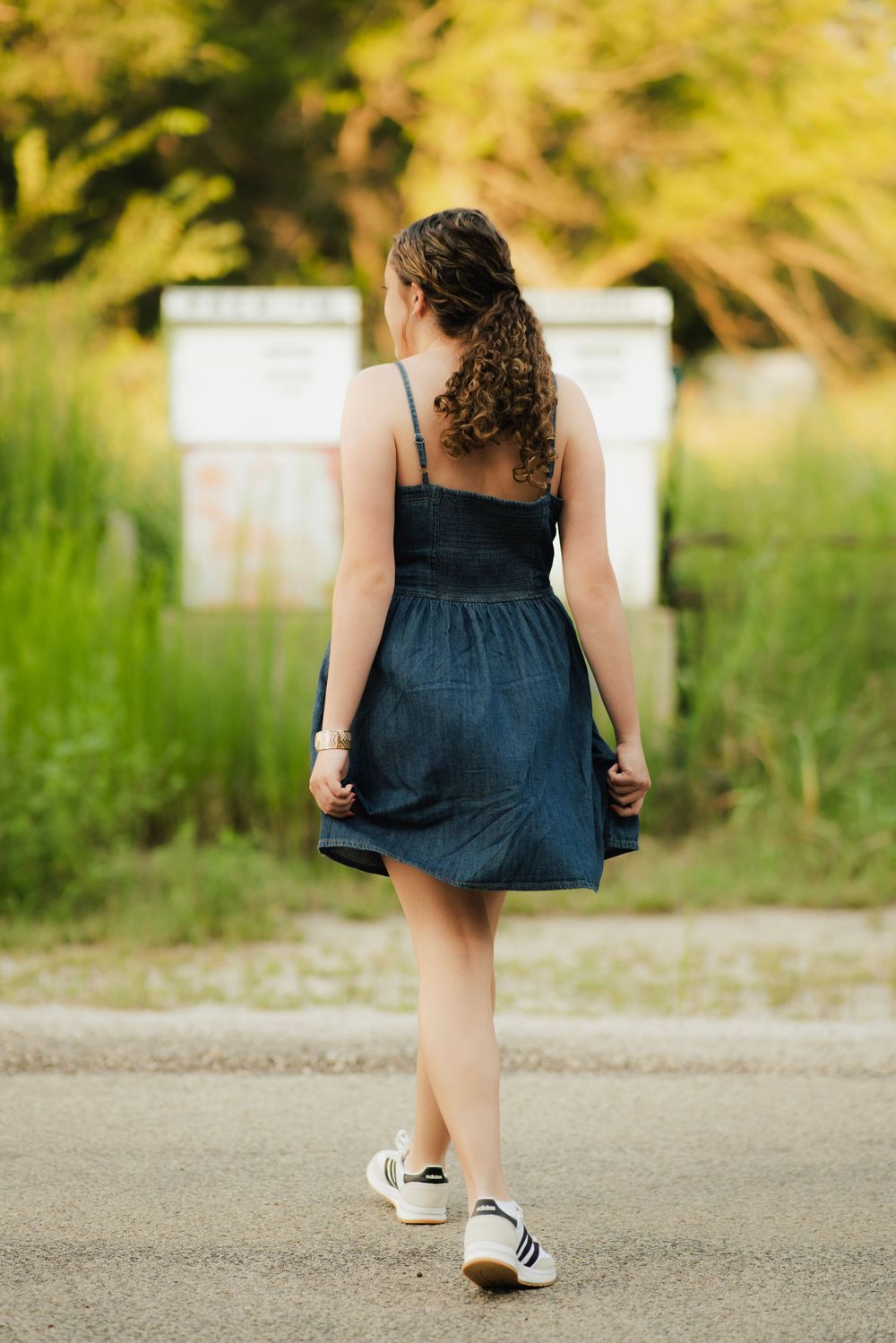 Young woman in navy sundress and white sneakers walking away on a path surrounded by lush greenery.