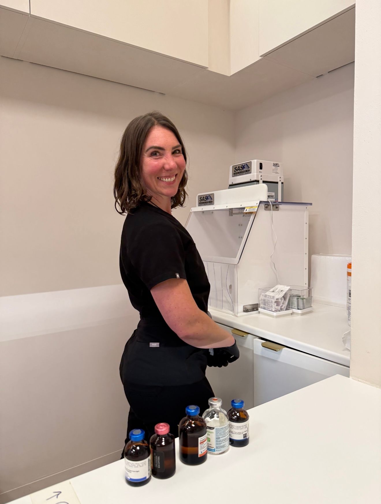 Smiling woman in black scrubs working at laboratory counter with colorful chemical bottles.