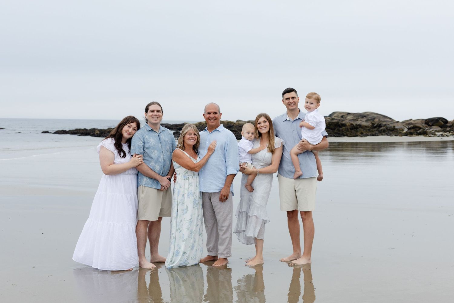 Family of seven poses on a misty beach, holding hands in a line, dressed in white and blue outfits.