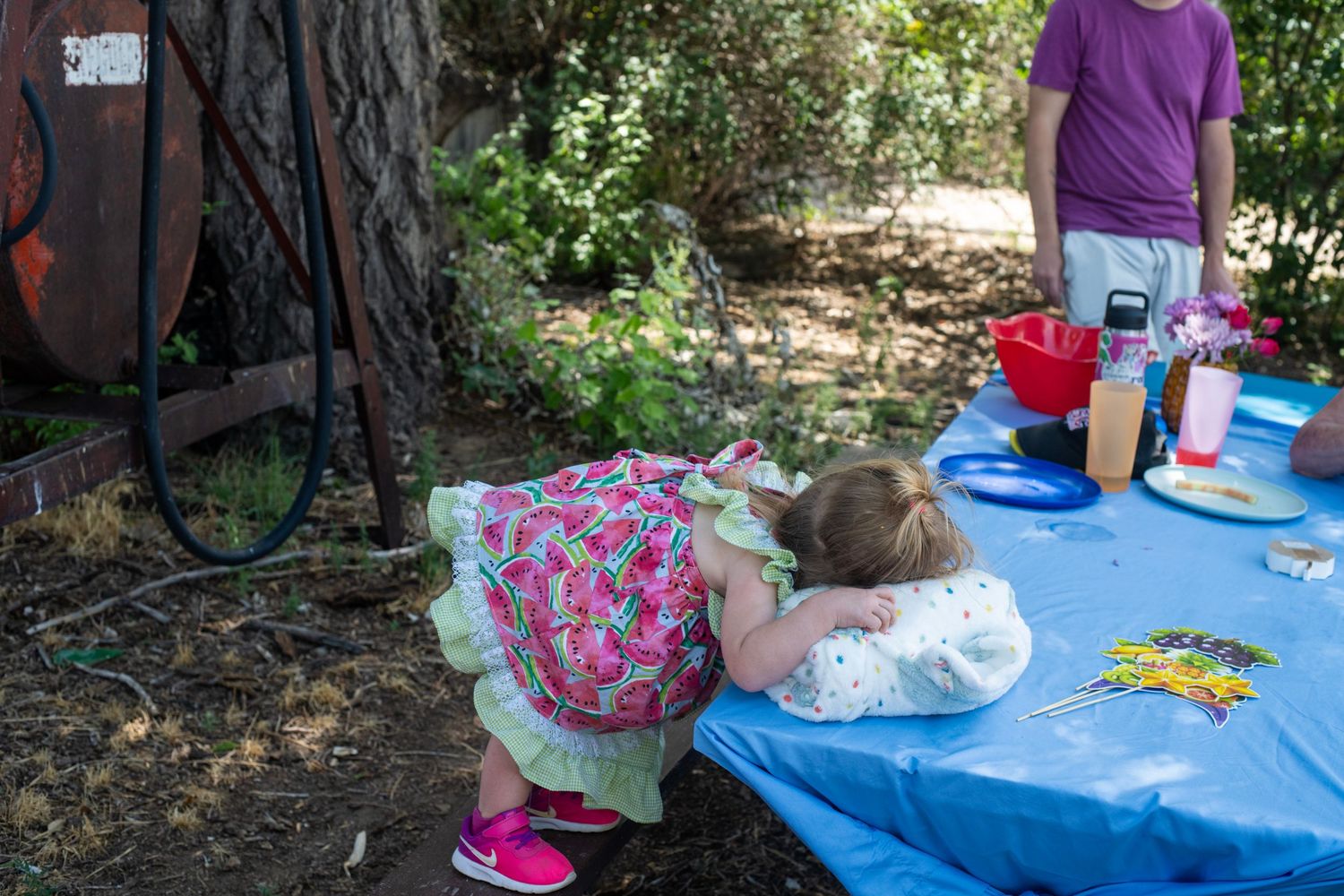 Young girl in floral dress leaning over blue tablecloth at outdoor birthday party with trees in background.