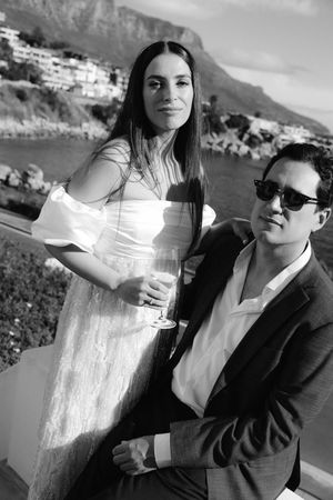 Black and white photo of an elegant couple at a scenic coastal venue, woman in off-shoulder dress holding champagne glass.