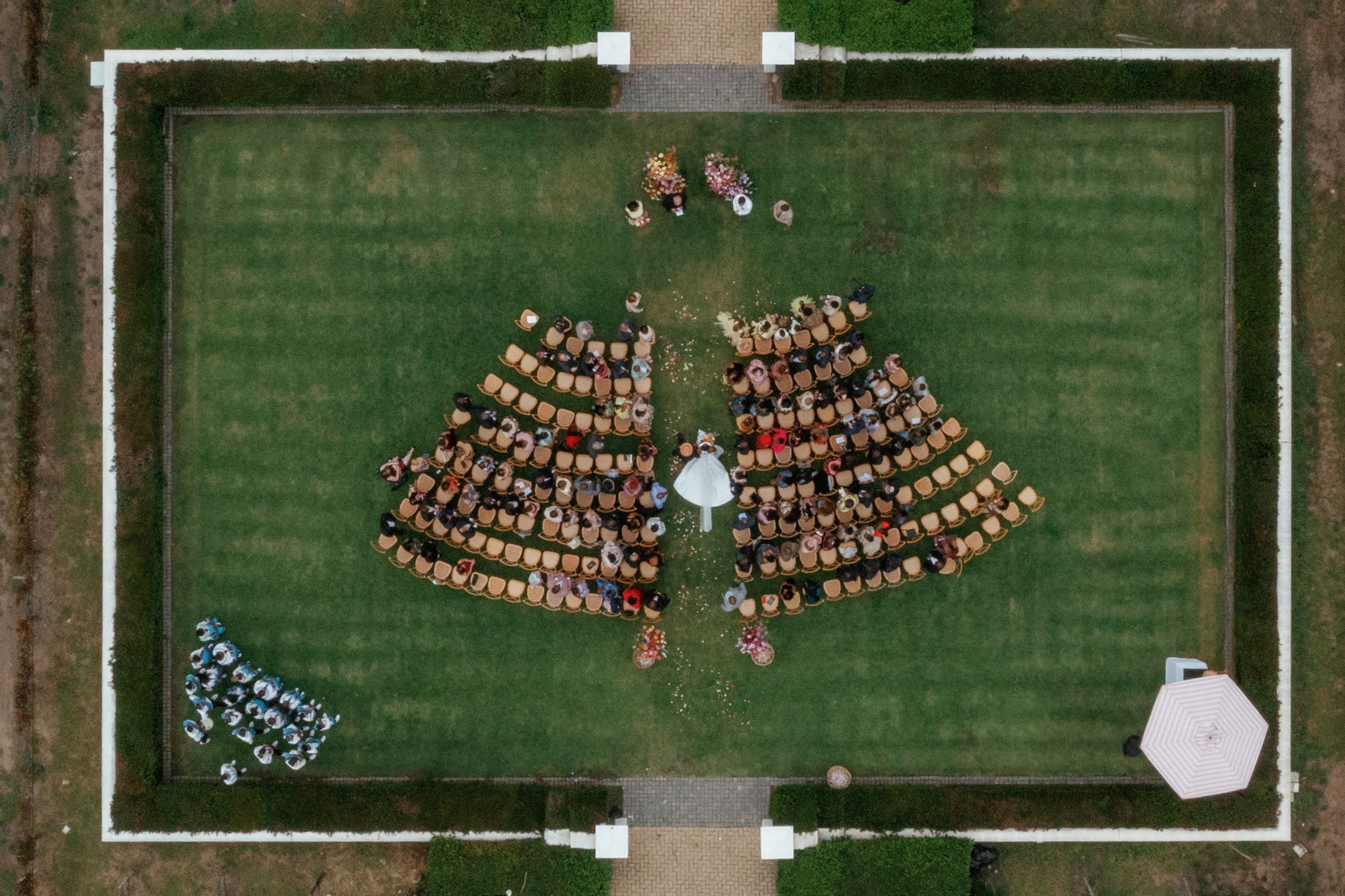 Aerial view of an outdoor wedding ceremony with guests seated in two curved rows on a green lawn.