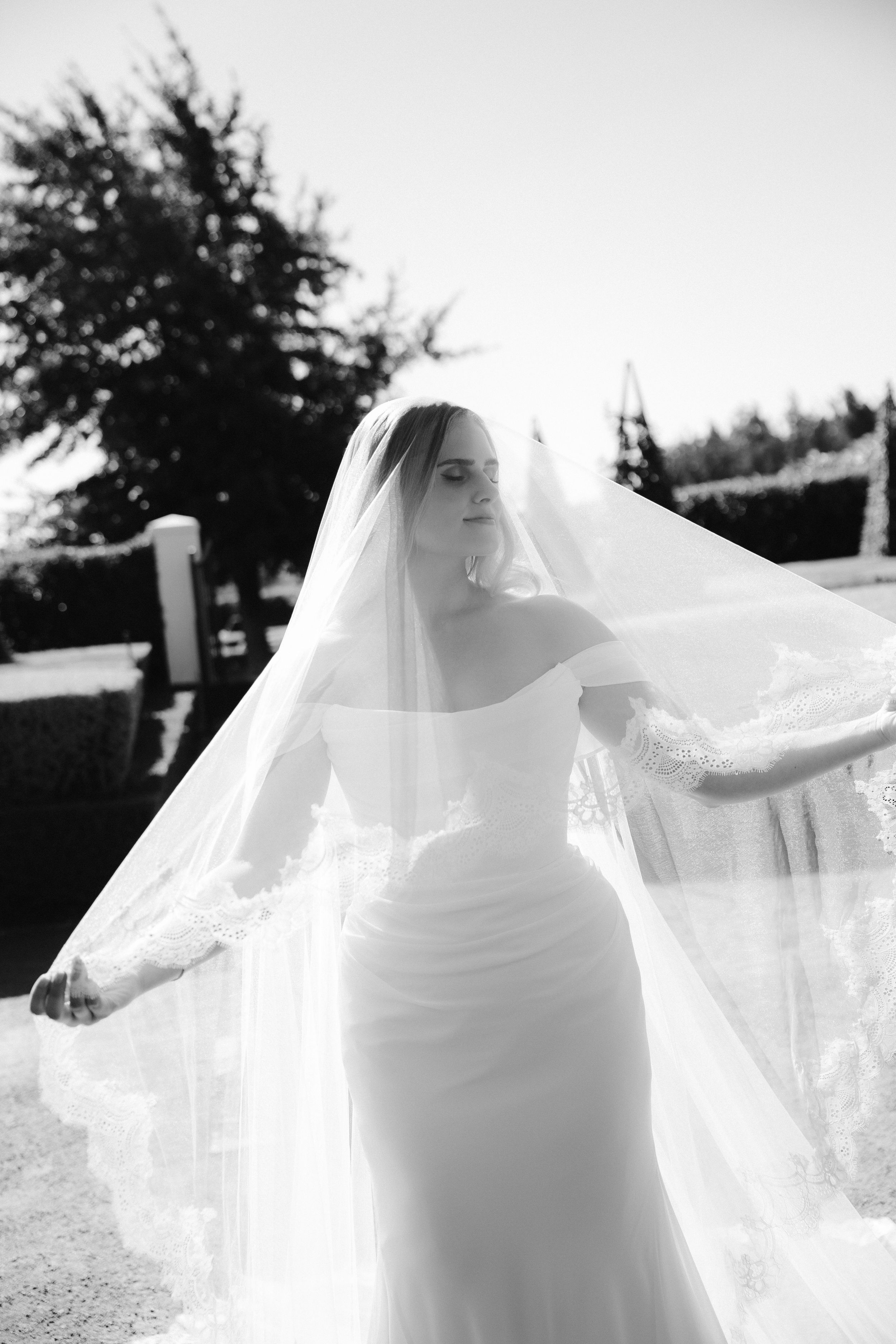 Black and white photo of a bride in an off-shoulder gown with a flowing lace-trimmed veil outdoors.