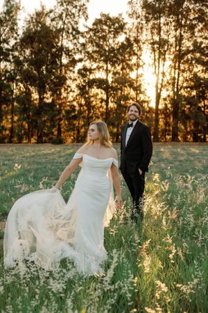 Bride in off-shoulder gown and groom in tuxedo walk through golden meadow at sunset with trees behind.
