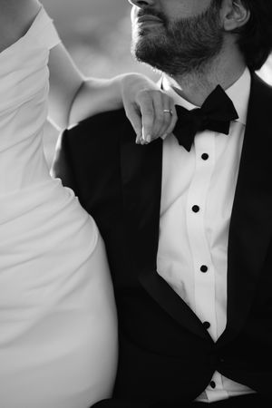 Black and white photo of a groom in a tuxedo with bow tie, bride's hand resting on his shoulder.