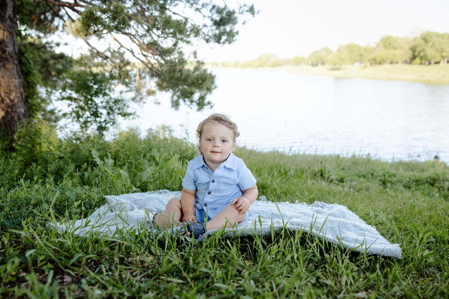 Baby boy sitting on a blanket in green grass near a lake, smiling outdoors in natural light.