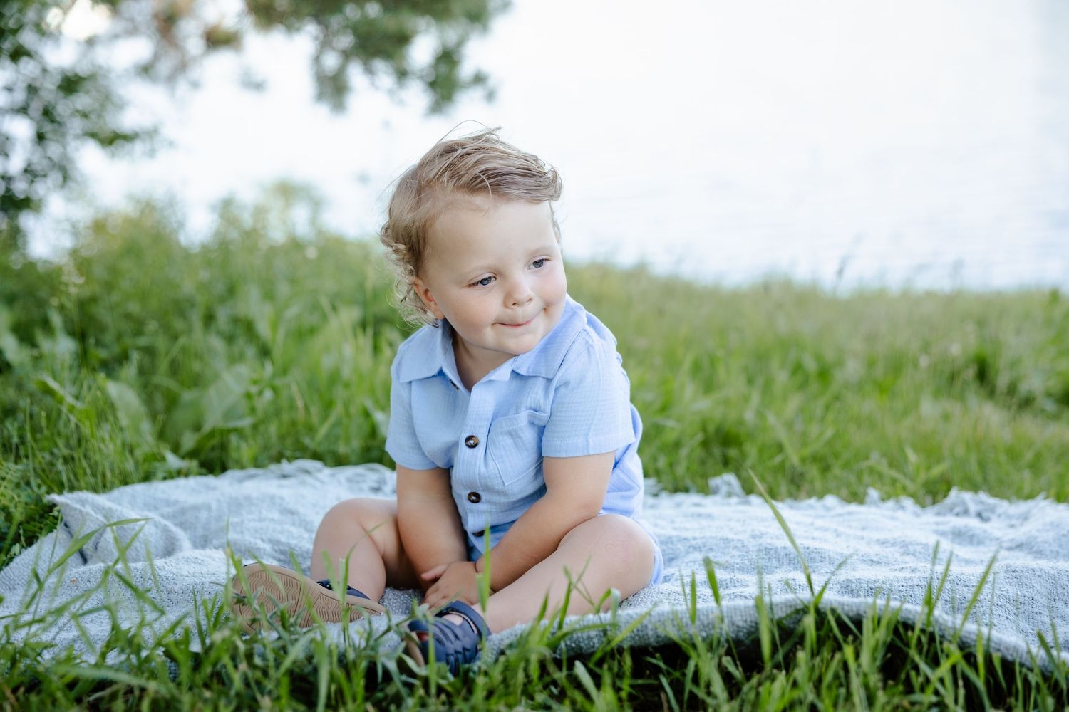 A young toddler boy in a blue shirt sits cross-legged on a blanket in lush green grass near water.