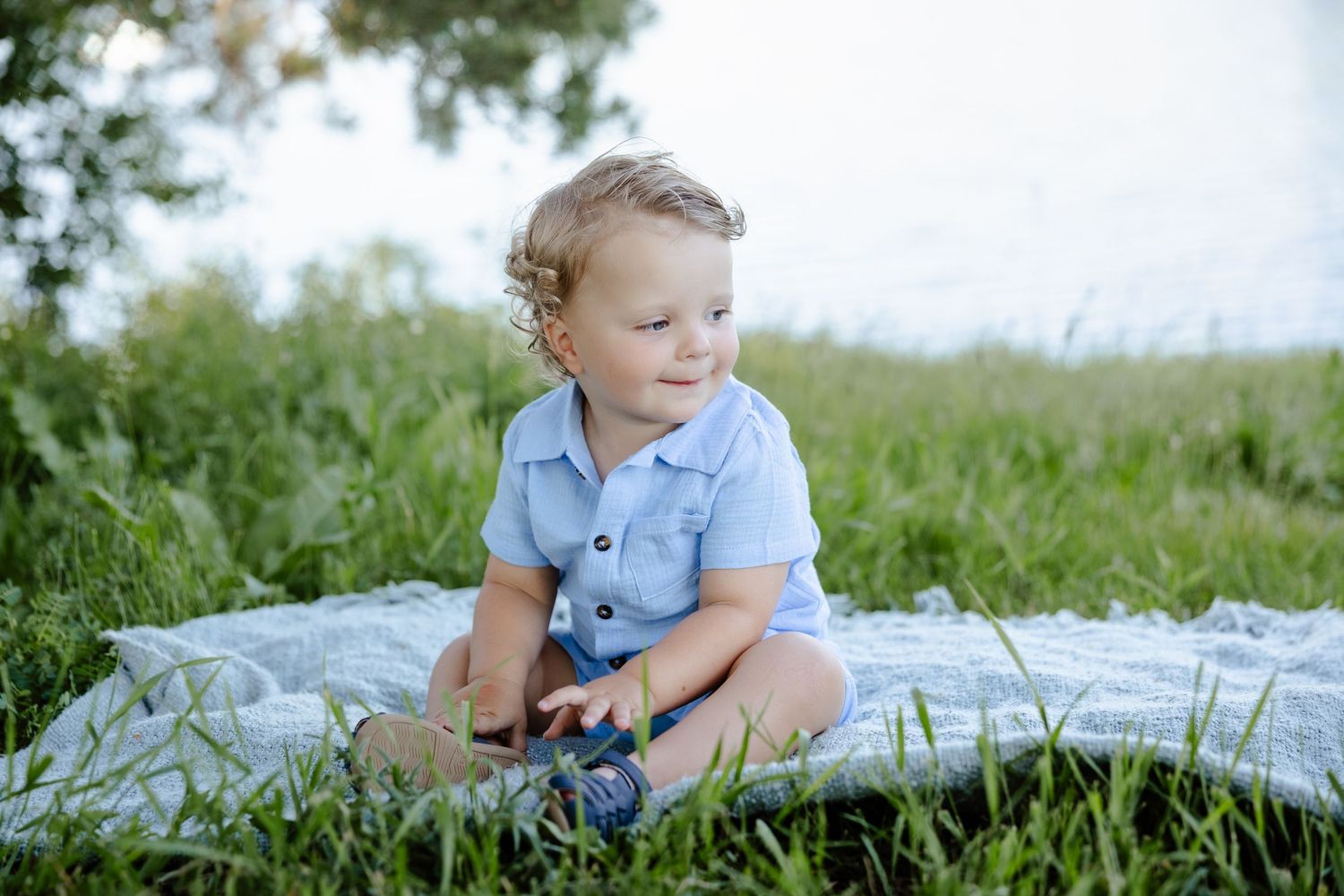 Curly-haired toddler boy in blue romper sitting on blanket in green grass near water.