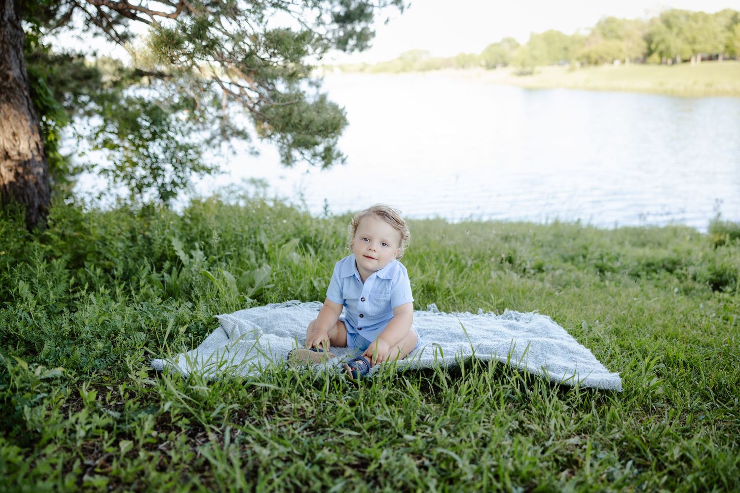 A smiling baby boy sits on a blanket in lush green grass near a serene lake on a sunny day.