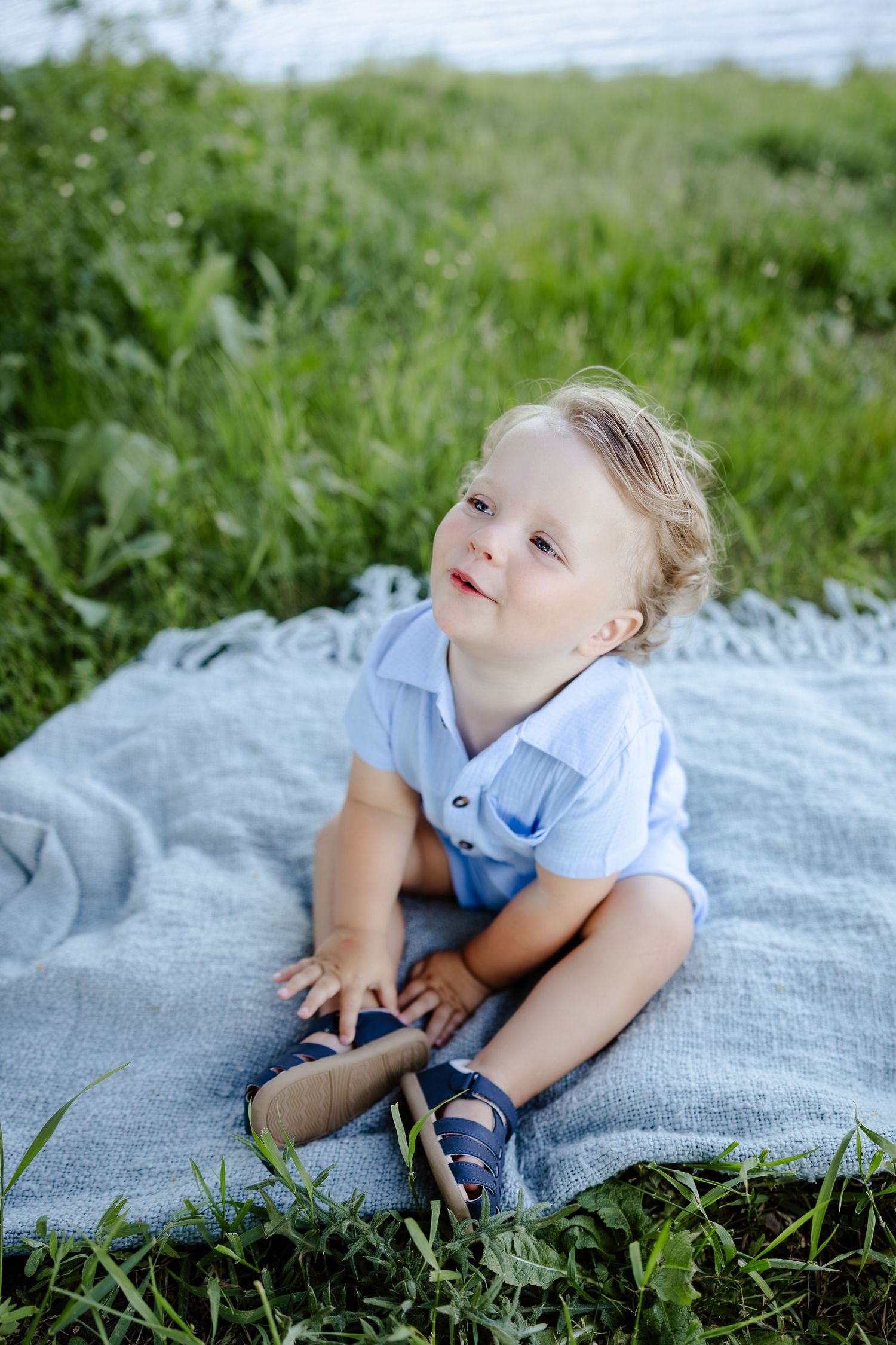 Cute toddler boy with curly blonde hair sitting on a blue blanket in green grass outdoors.