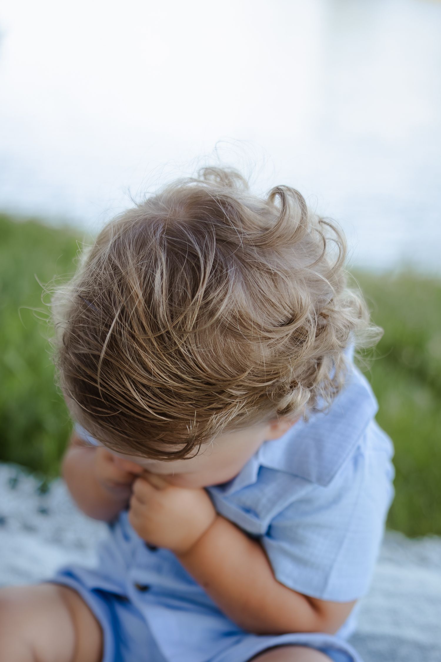 Toddler with curly blonde hair wearing a blue shirt, sitting outdoors near green grass, looking down.