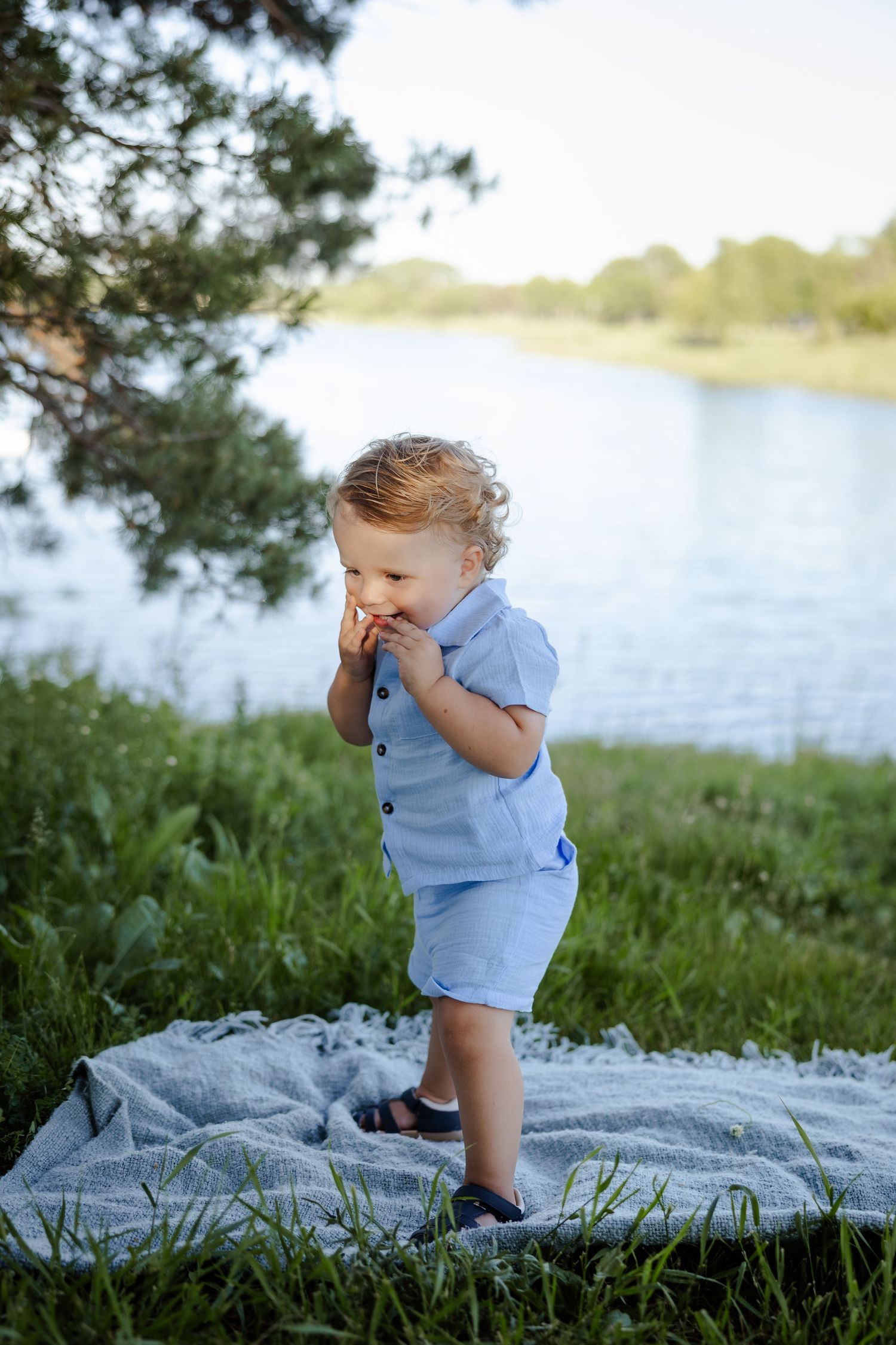 Curly-haired toddler in blue outfit standing on blanket by lake eating snack outdoors.