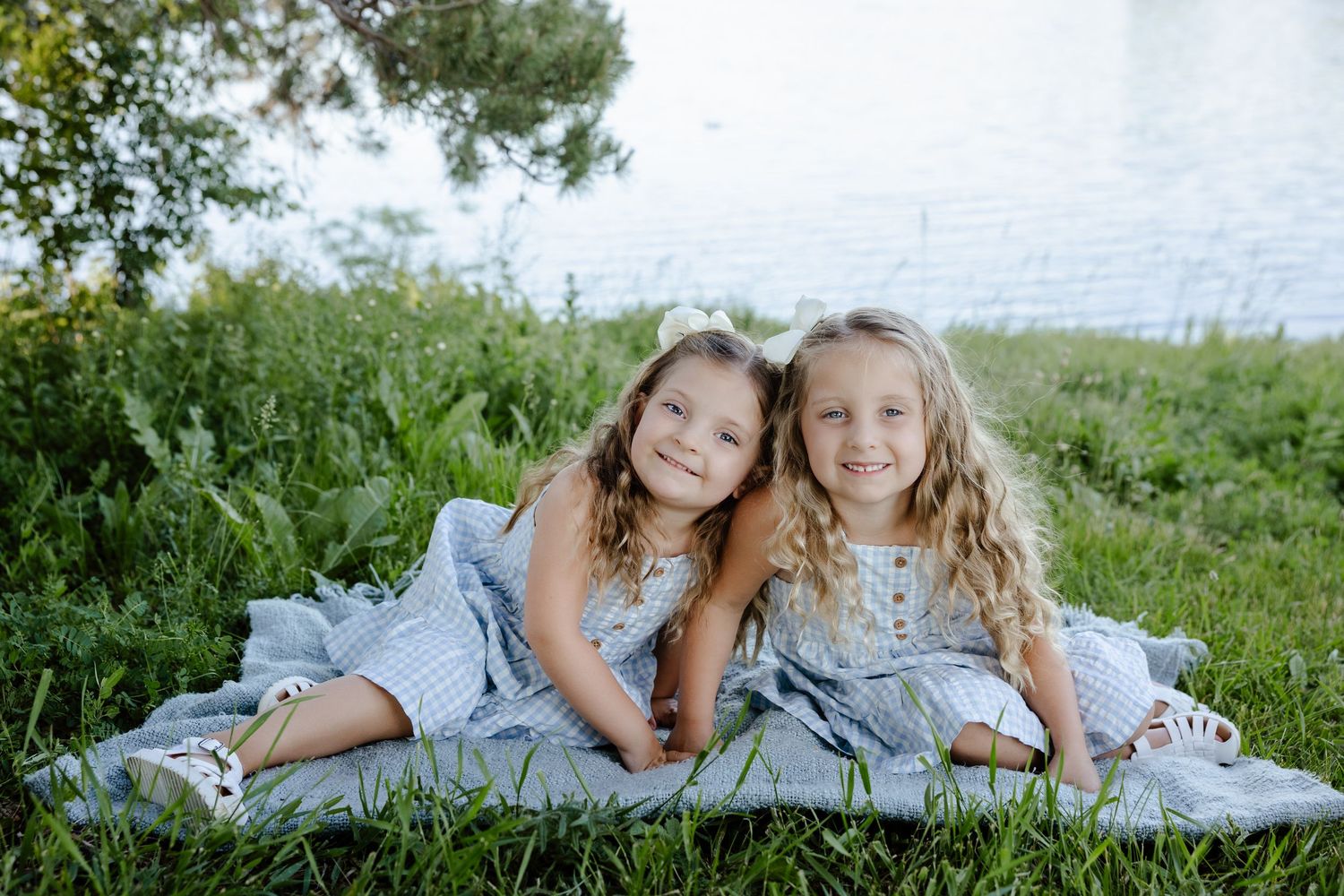 Two young girls in matching blue dresses smiling together on a blanket in green grass near a lake.