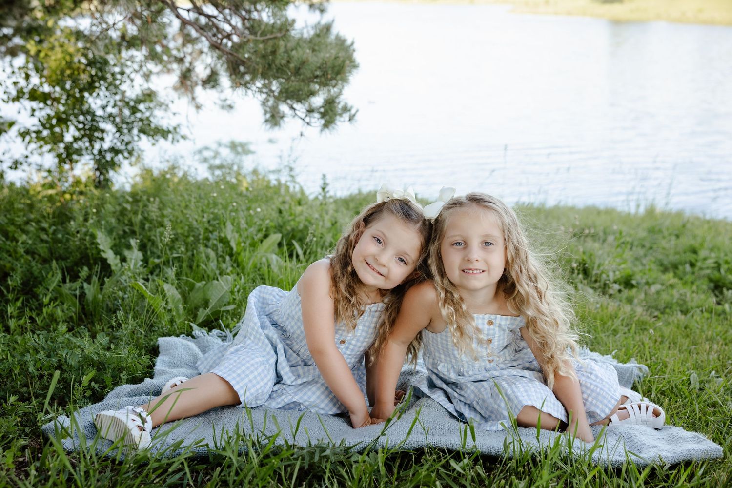 Two young girls in matching blue dresses sitting together on a blanket by a lake surrounded by green grass.