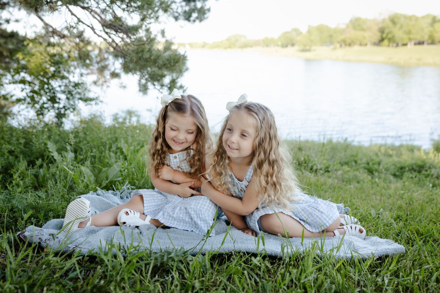 Two young girls in matching blue dresses sit together on a blanket by a peaceful lake surrounded by green grass.