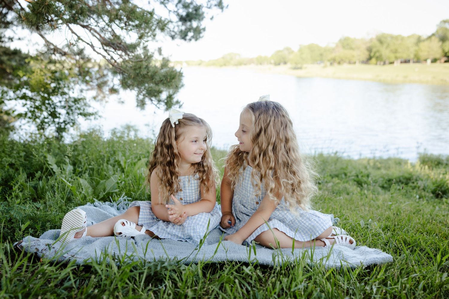 Two young girls in matching gray dresses sit on a blanket by a river, smiling at each other outdoors.
