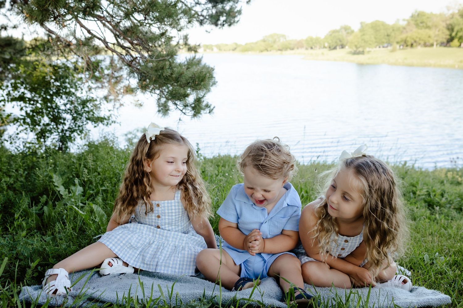 Three young children sitting together on a blanket by a serene lake surrounded by lush green trees.