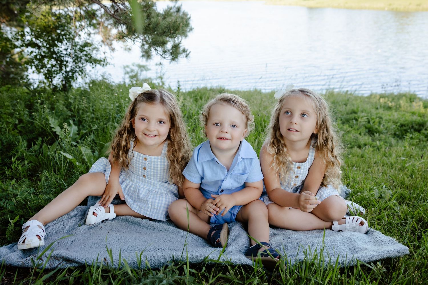 Three young siblings with curly hair sitting together on blanket in grass by lake.