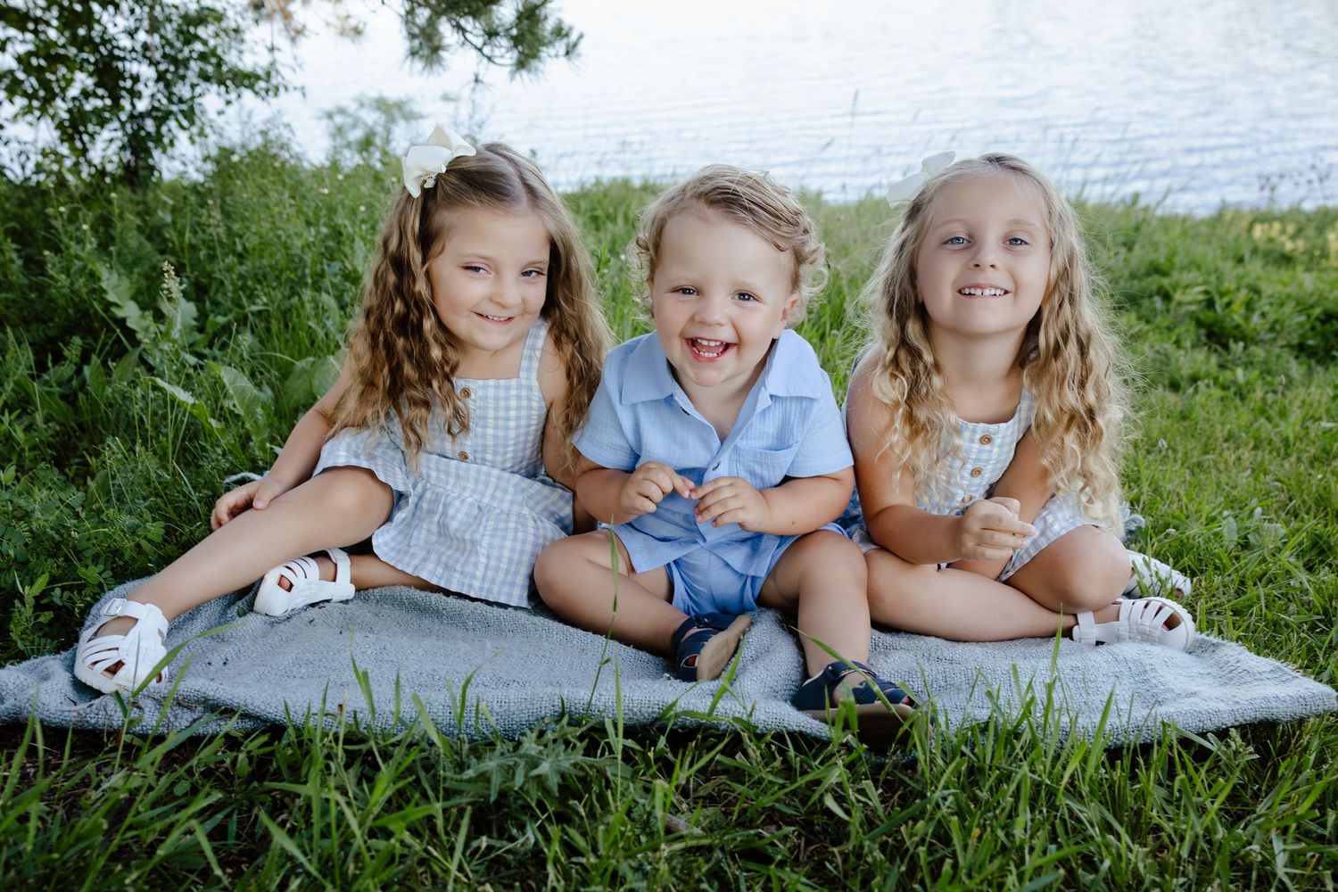 Three happy children sitting on a blanket in green grass near a lake, smiling outdoors in casual summer outfits.