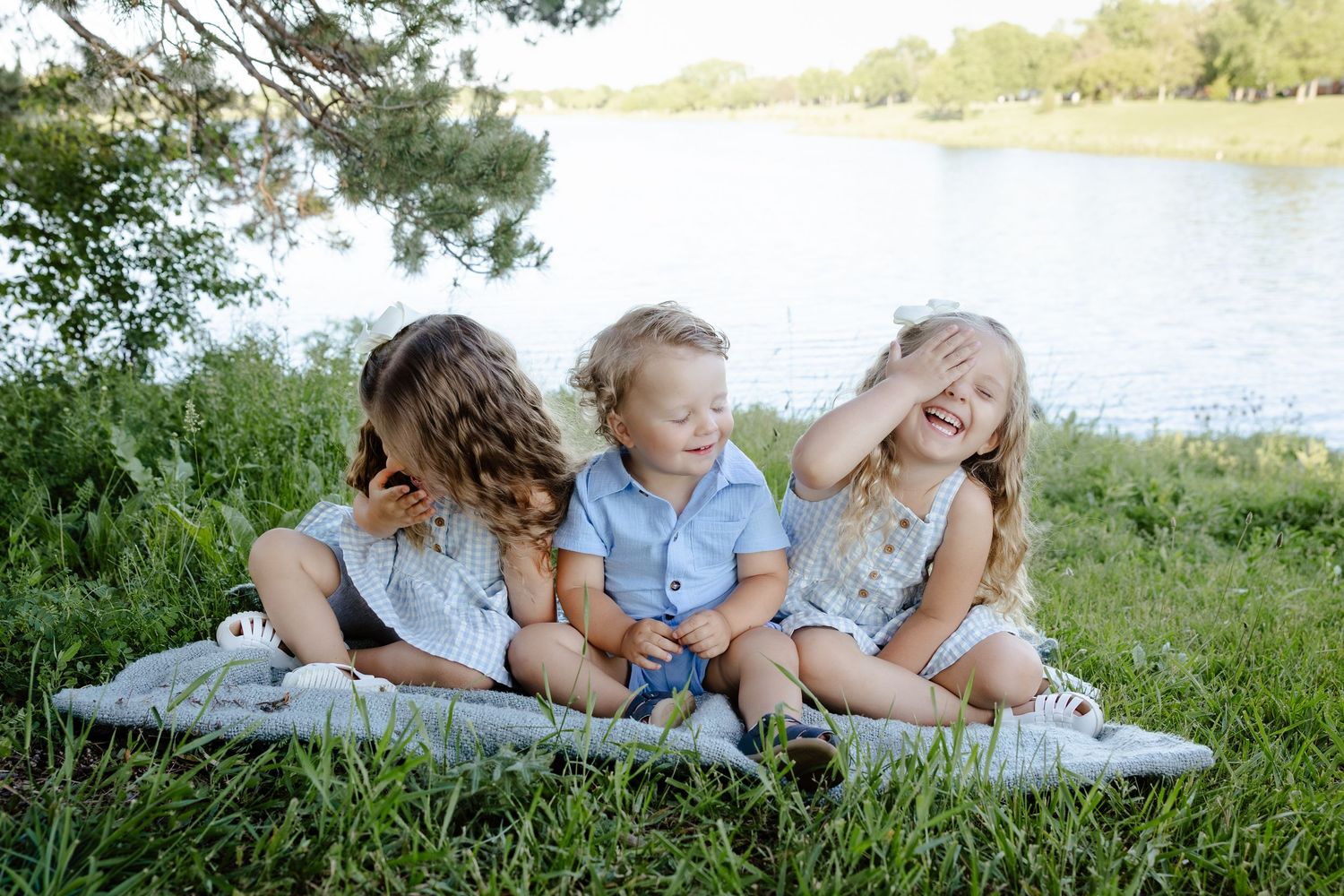 Three young siblings sit together on a blanket by a river, laughing and playing outdoors in summer.