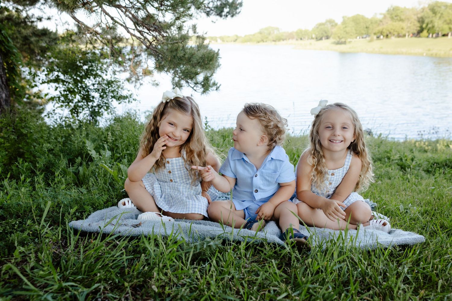 Three young siblings smile and sit together on a blanket by a peaceful lake surrounded by lush green grass.