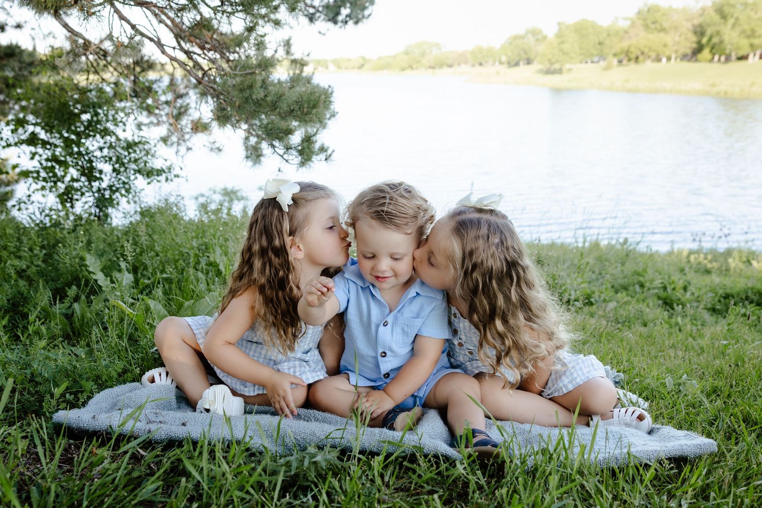 Two sisters give their little brother sweet kisses on the cheeks while sitting on a blanket near a lake.