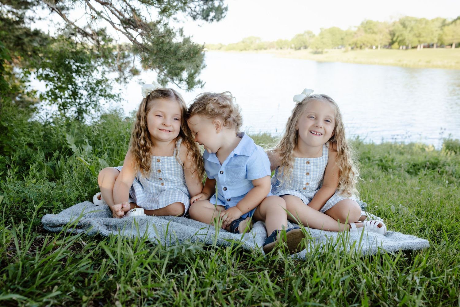 Three young children sit together on a blanket by a lake, smiling and enjoying a sunny outdoor day.