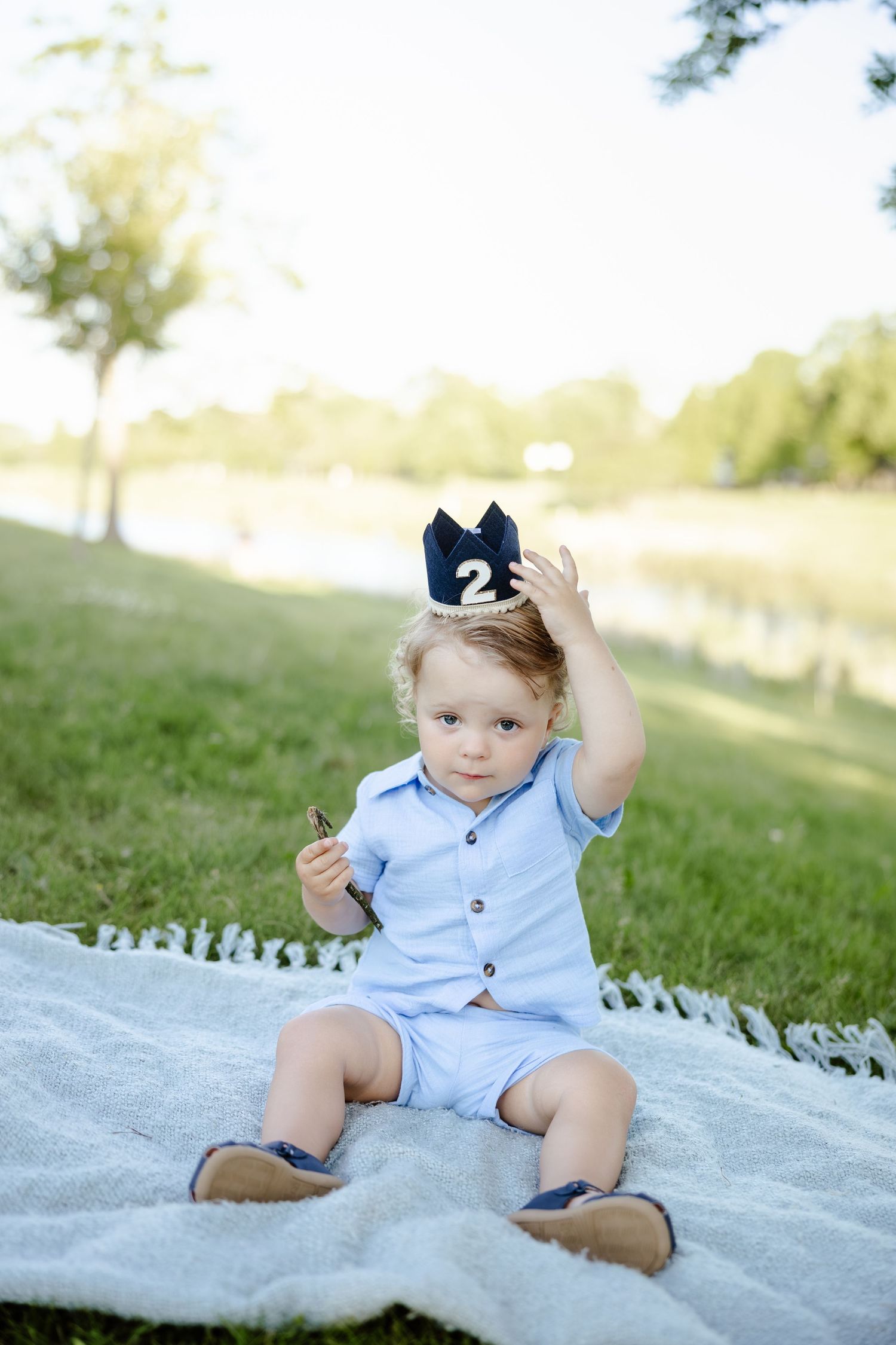 Toddler boy in blue outfit wearing a number 2 crown sitting on blanket outdoors for birthday photos.