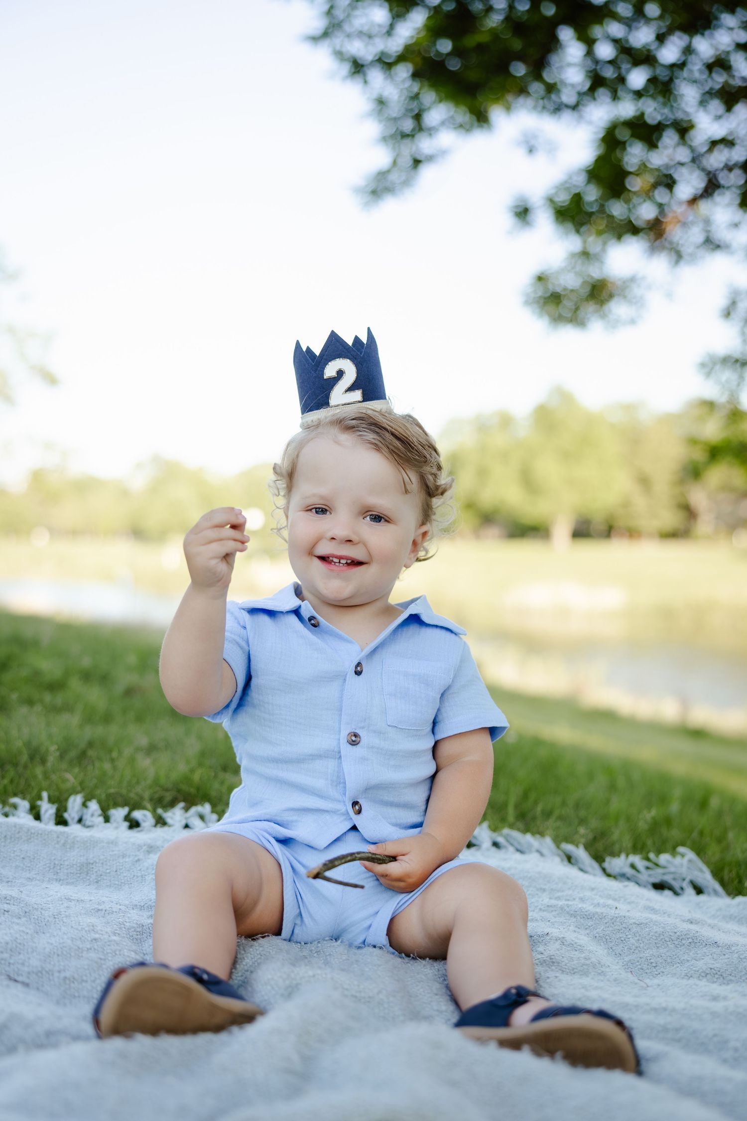 A smiling toddler boy in a blue outfit sits on a blanket outdoors wearing a number 2 birthday crown.