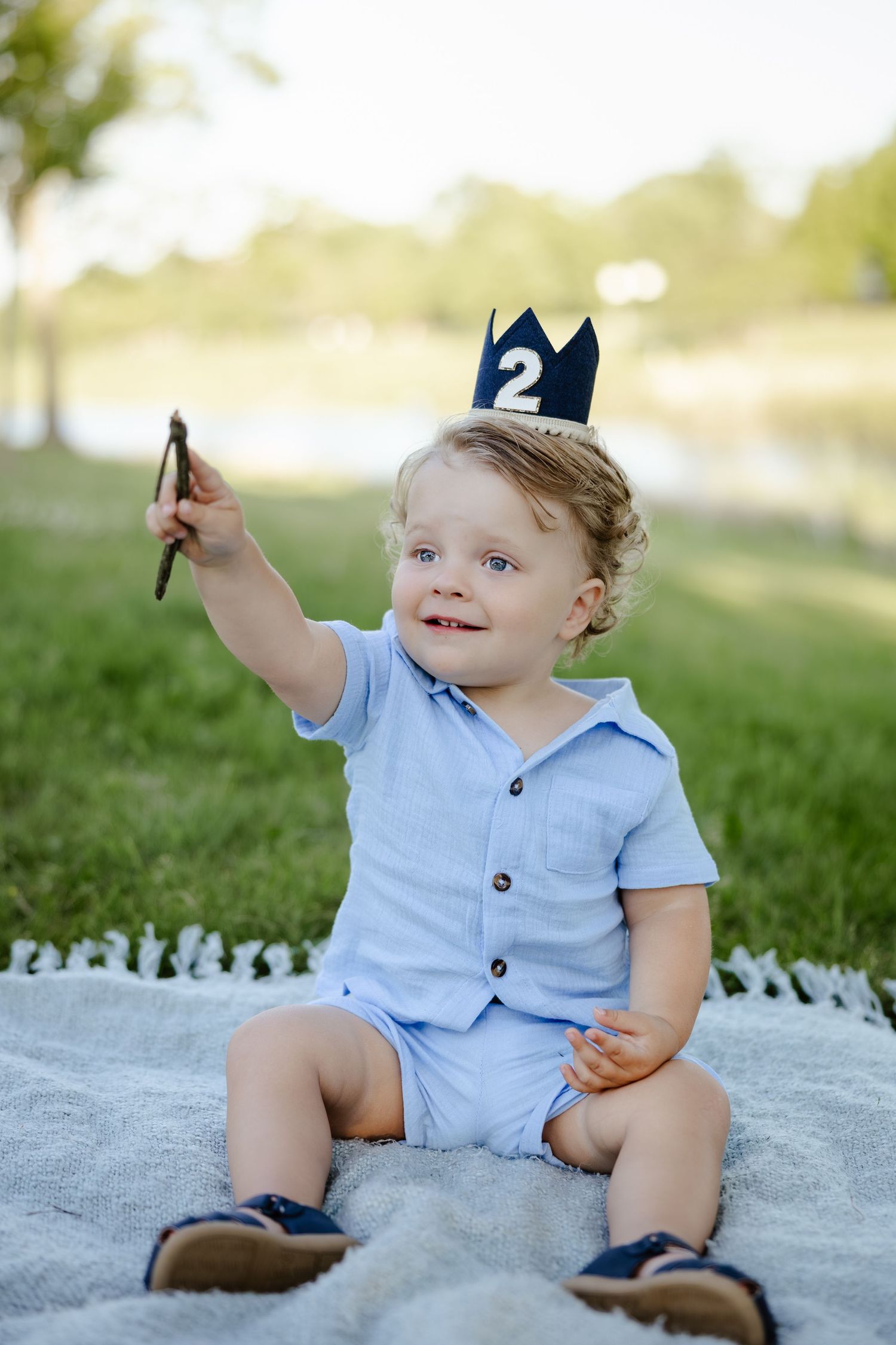 Toddler boy in blue outfit wearing a number 2 crown sits on blanket outdoors celebrating his 2nd birthday.
