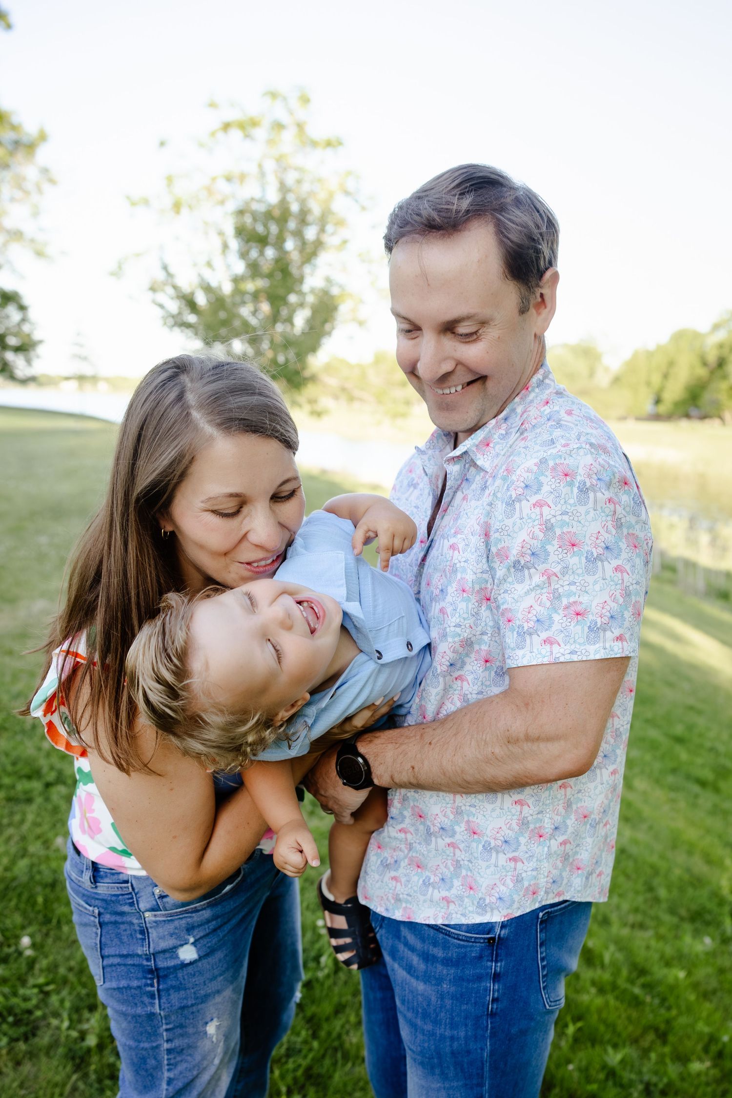 Happy family with parents holding laughing toddler boy in sunny green park.