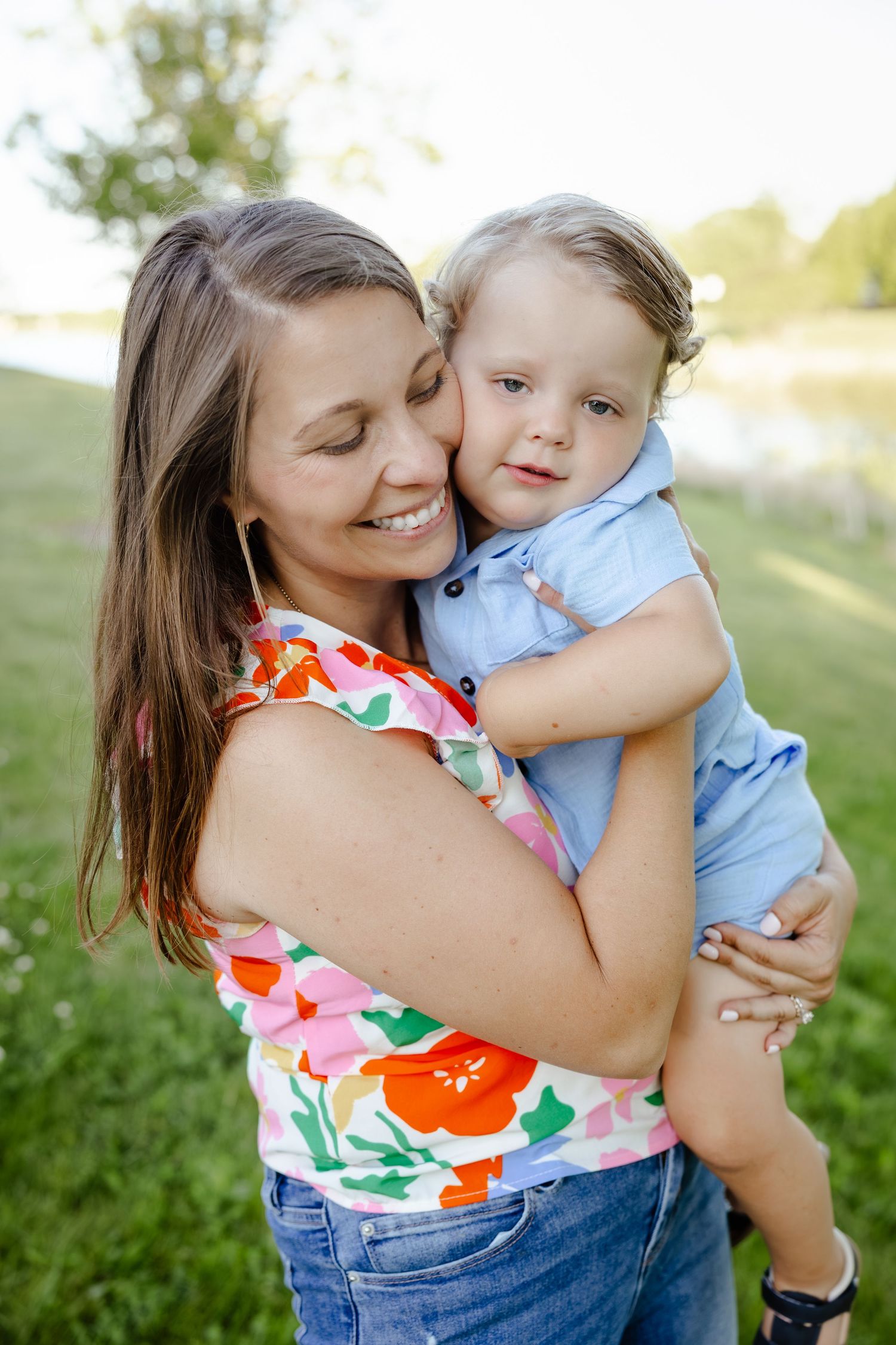 Smiling mother in floral top hugging her toddler boy outdoors in a green park setting.