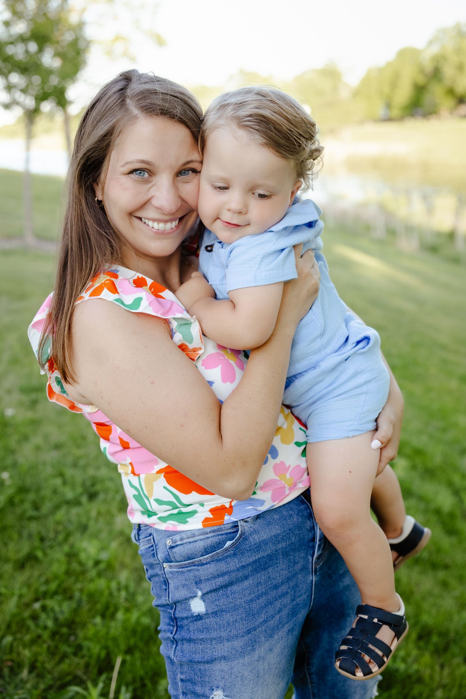 Smiling mother holding young toddler boy outdoors in green park setting, wearing colorful floral top and jeans.