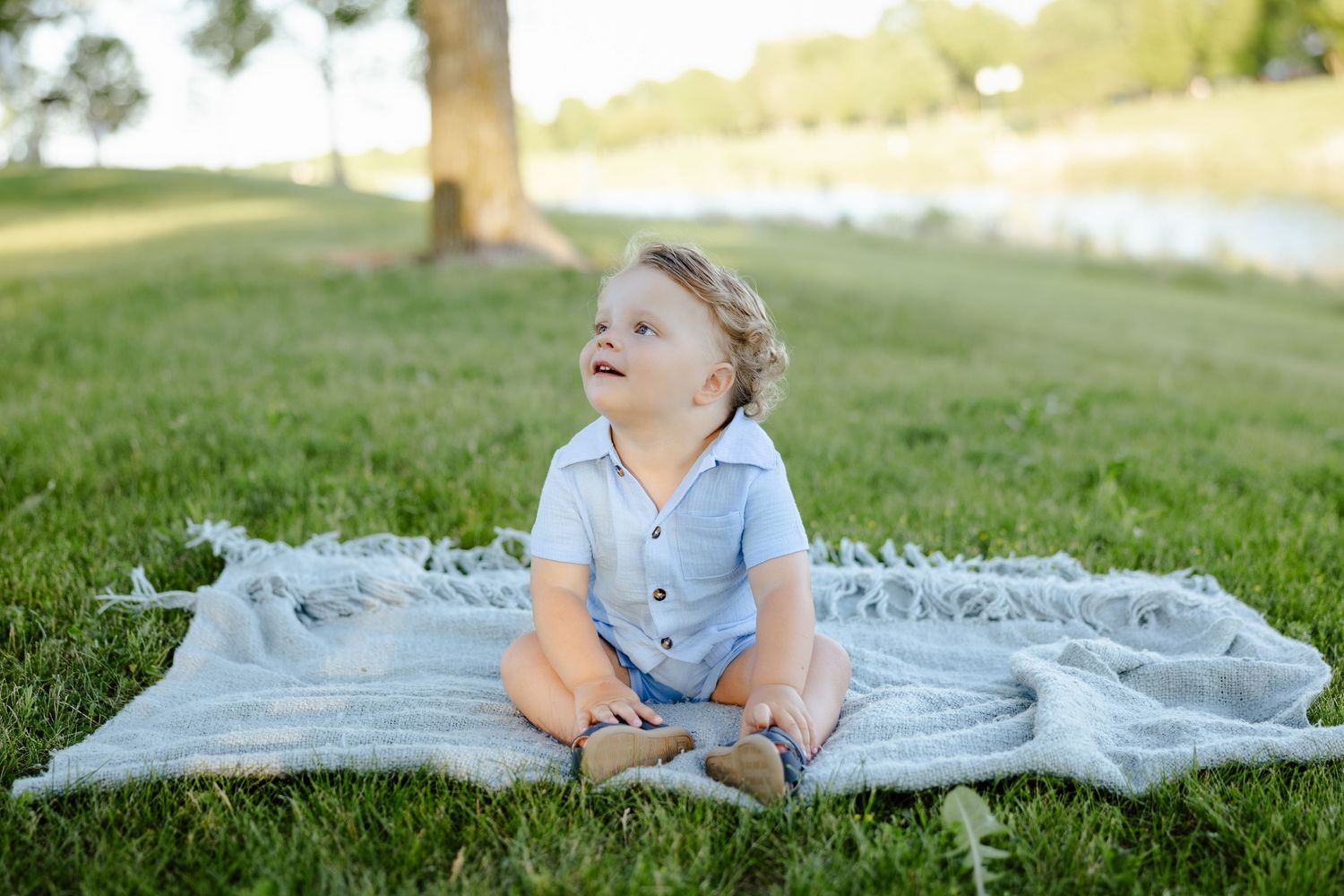 Adorable baby boy sitting on blanket in lush green park near lake.