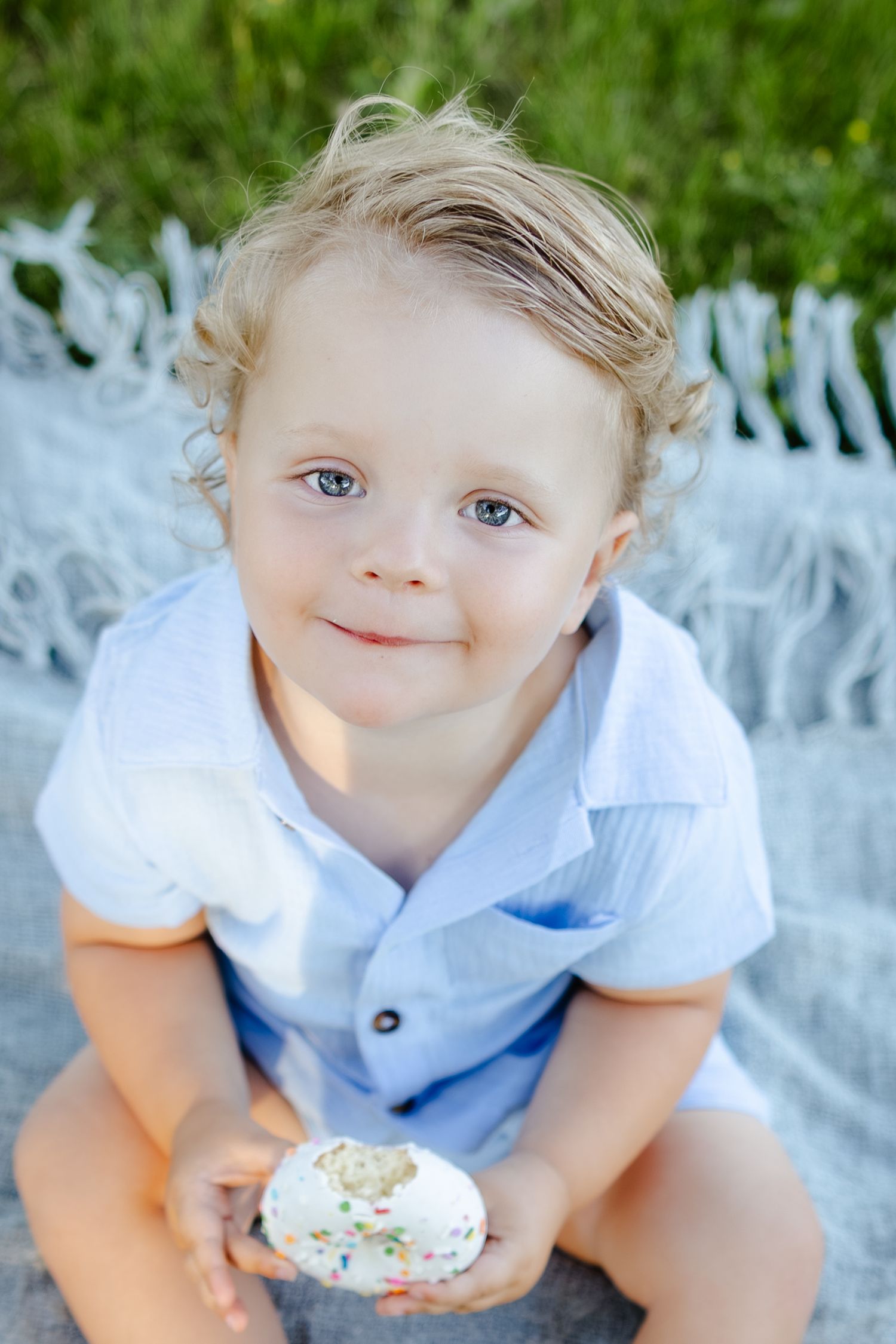Smiling toddler boy with curly blonde hair holding a frosted cupcake with sprinkles while sitting on a blanket outdoors.