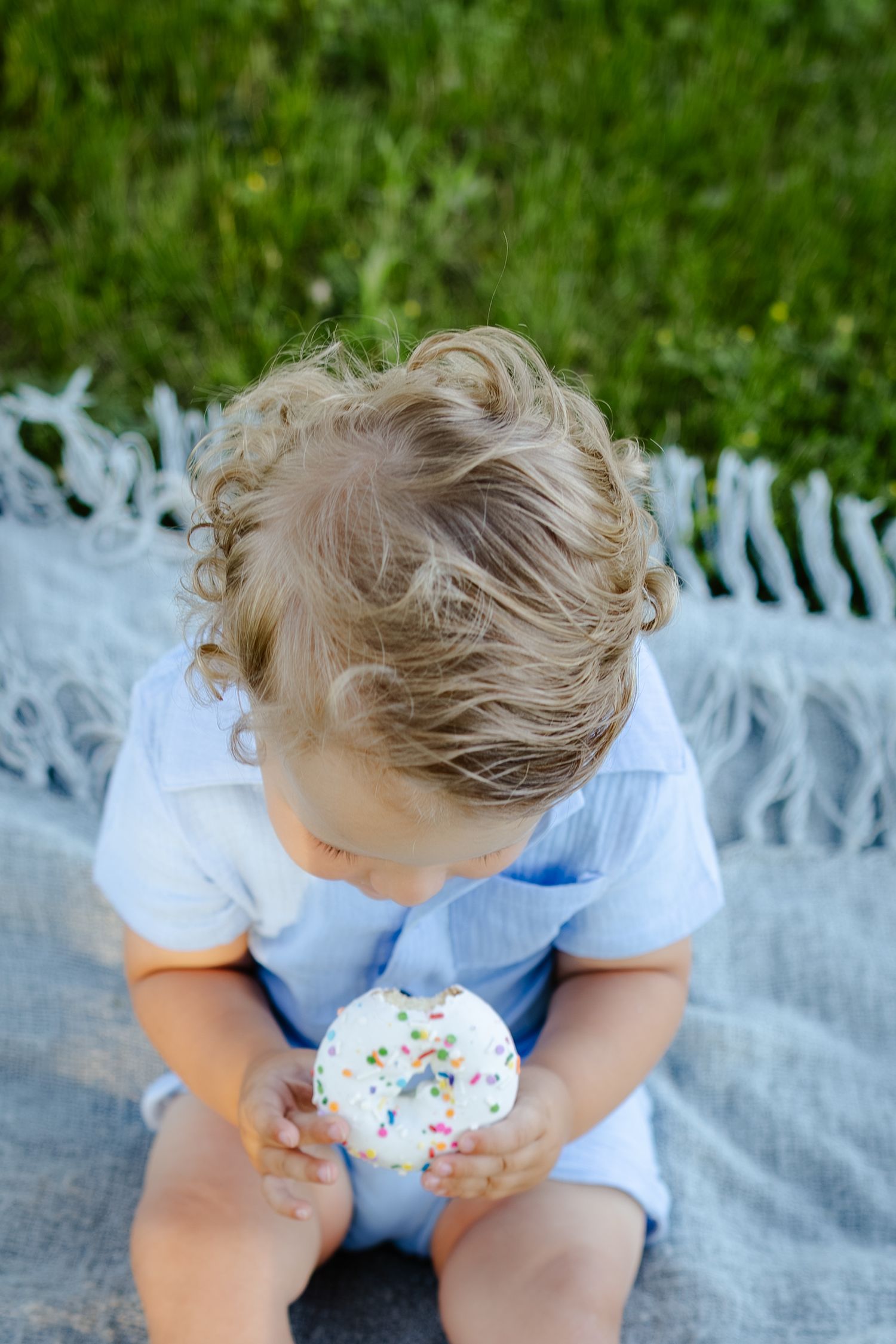 Curly-haired toddler boy in a blue shirt looking down at a white frosted cupcake with colorful sprinkles on a gray blanket.