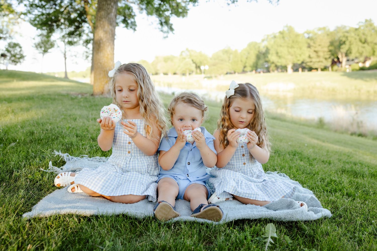 Three young children sitting on a blanket outdoors eating snacks near a pond on a sunny day.