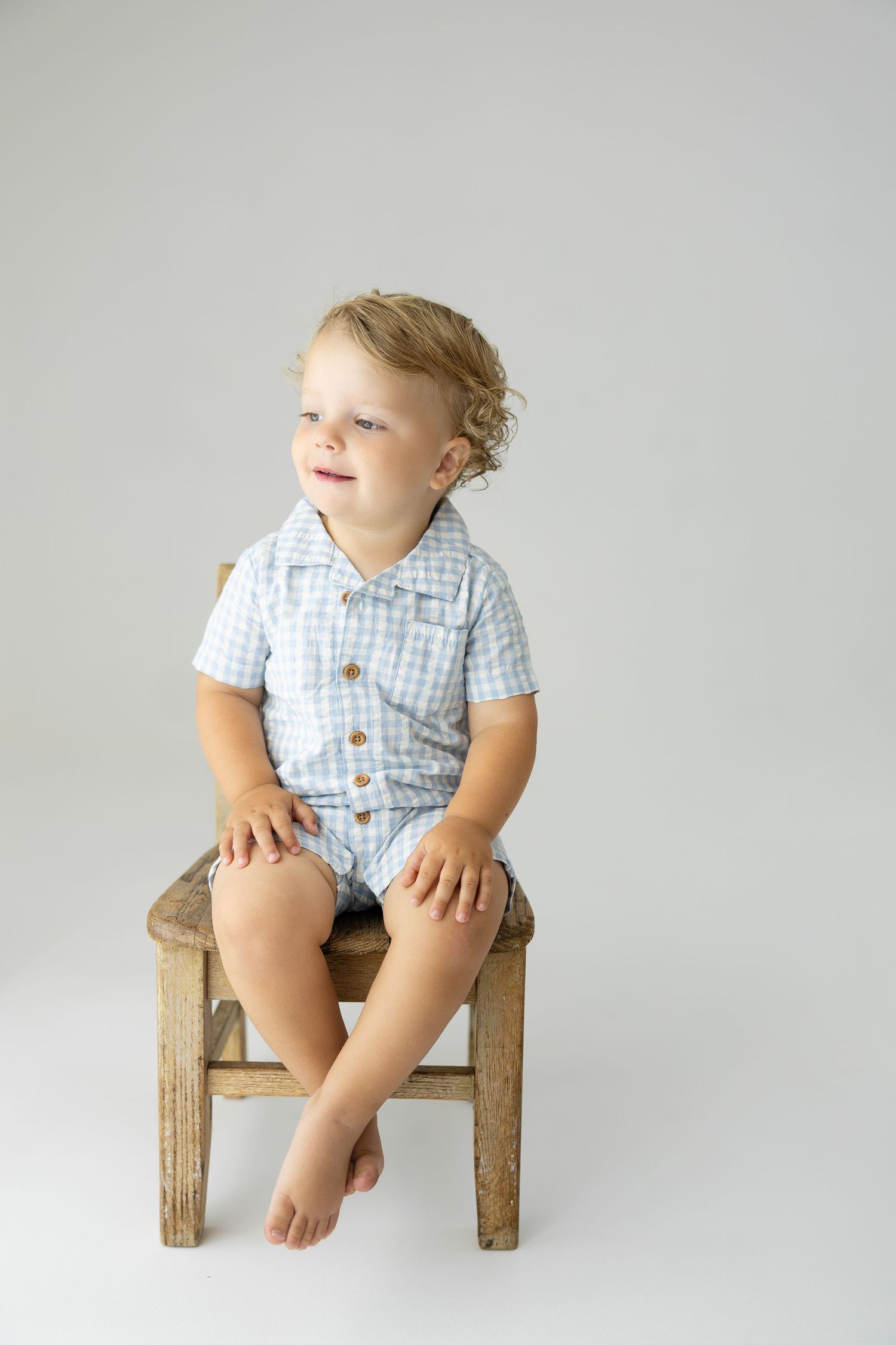 Cute blond toddler boy sitting on rustic wooden stool wearing light blue checkered romper on white background.