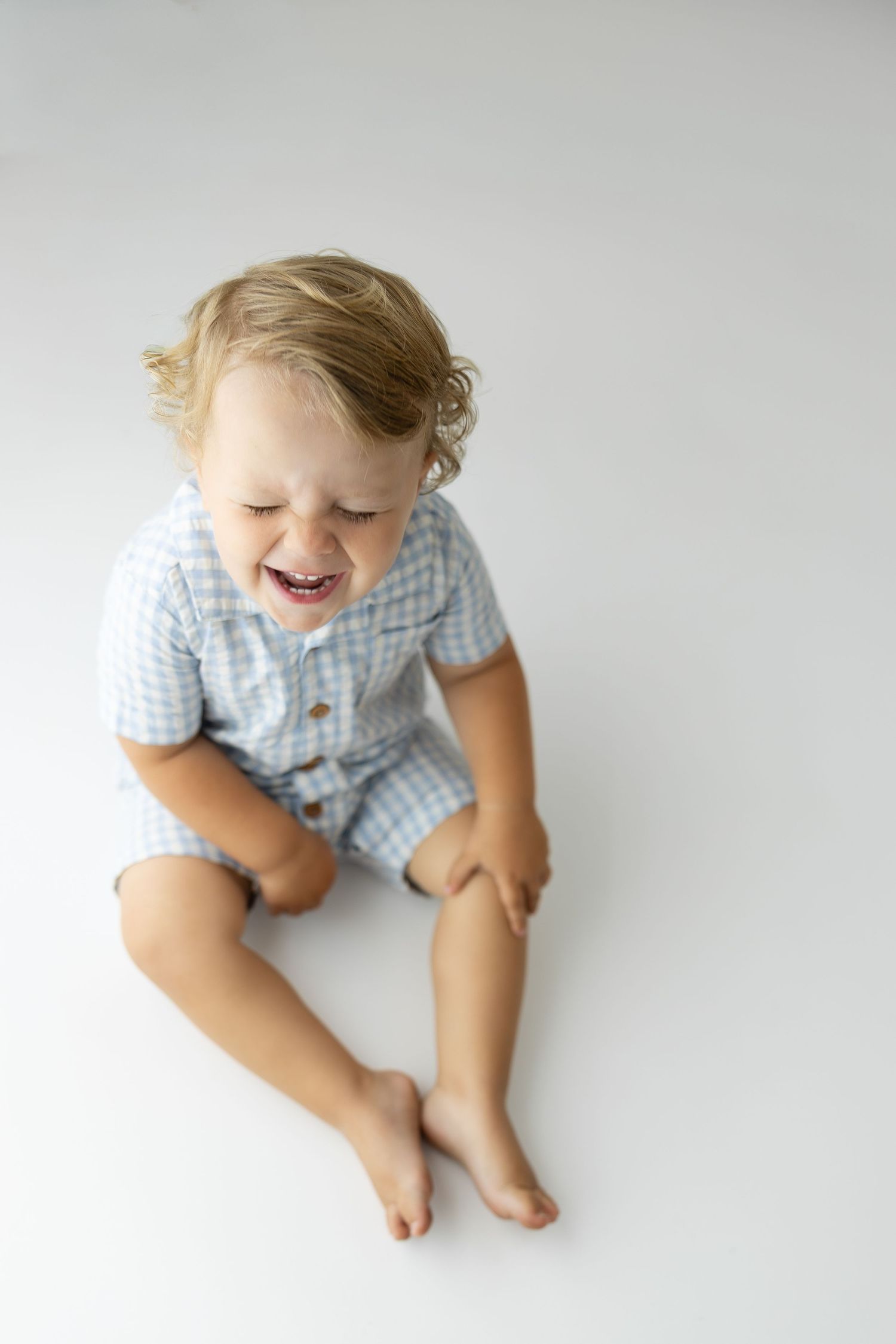 Laughing toddler boy with curly blonde hair sitting barefoot on white floor wearing light blue gingham romper.