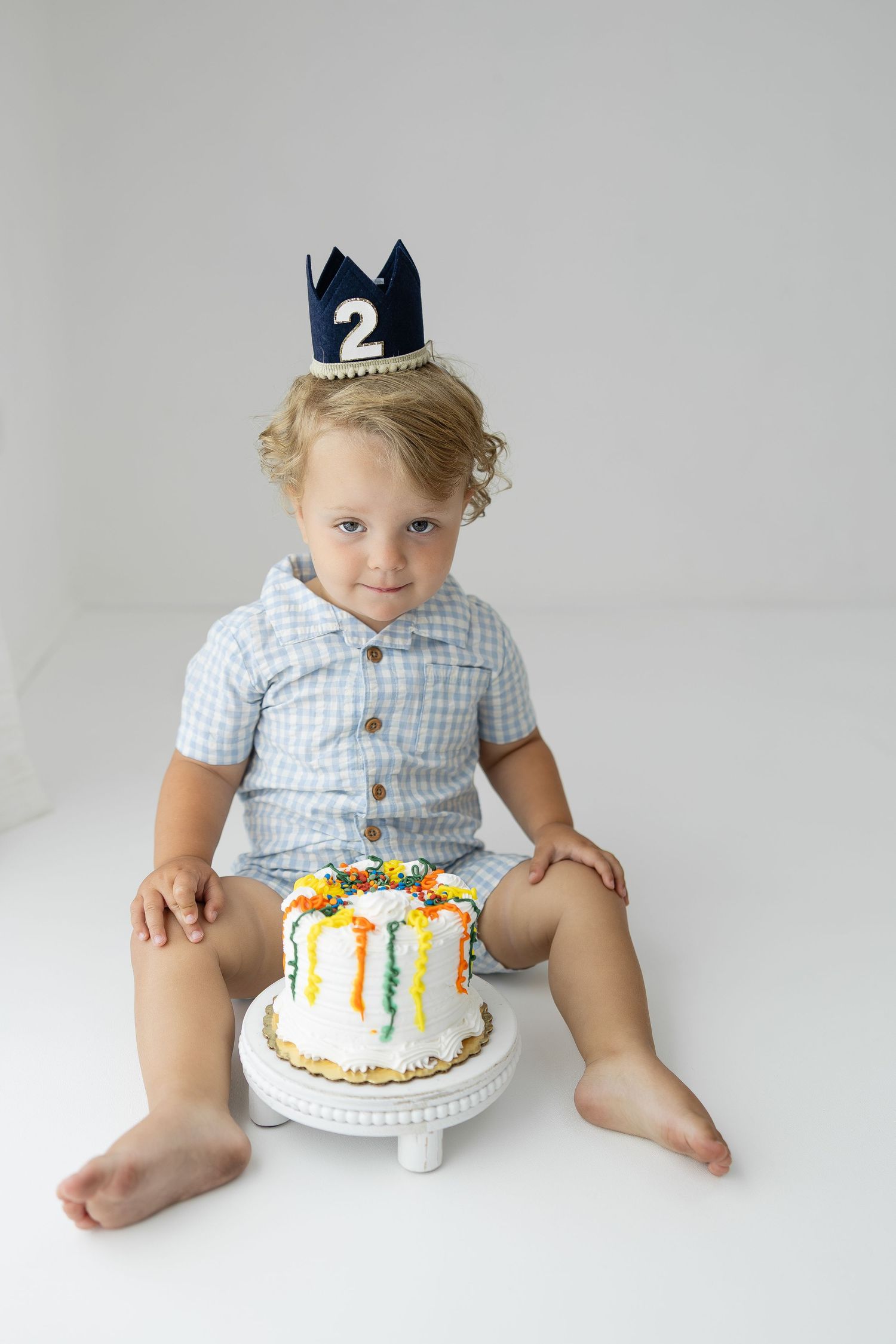 Blond toddler boy wearing number 2 crown sitting with colorful birthday smash cake on white background.